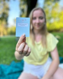 Woman outdoors holding a light blue The Well-being Game card toward the camera while sitting on a picnic blanket.