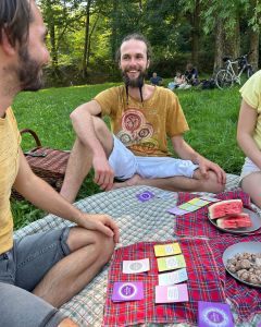 Two people sitting on blankets in a park with The Well-being Game cards, watermelon, and nuts placed in the center.