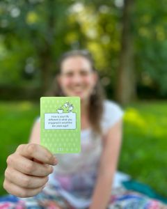 Woman smiling and holding up a green The Well-being Game card toward the camera during an outdoor picnic.