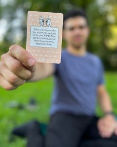 Man holding up a The Well-being Game card toward the camera while sitting on the grass during an outdoor gathering.