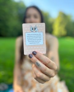 Woman holding a The Well-being Game card close to the camera during an outdoor picnic gathering.