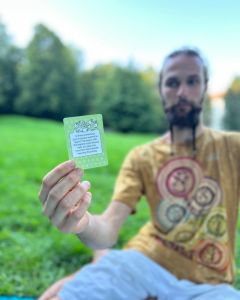 Man sitting on the grass holding up a The Well-being Game card toward the camera during an outdoor gathering.