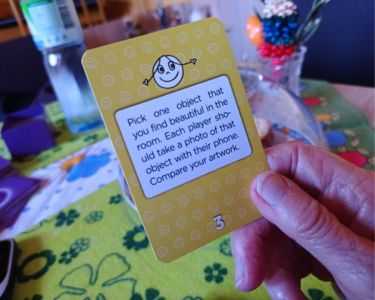 Hand holding a yellow Joy Card from The Well-being Game above a patterned tablecloth.