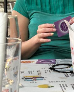 Person in a green top holding a purple card from The Well-being Game at a café table with drinks and glasses.