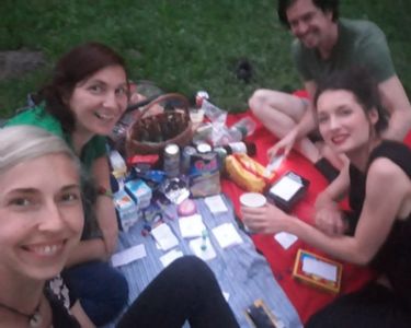 Group of four people sitting on picnic blankets in a park, with snacks and The Well-being Game cards laid out in front of them.