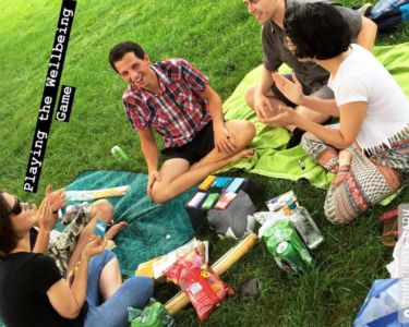 Group of people sitting on blankets in the grass, talking and playing The Well-being Game with snacks placed in the center.