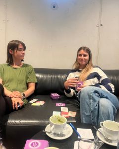 Two women sitting on a couch with The Well-being Game cards and drinks on the table in front of them.