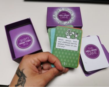 Hand holding several The Well-being Game cards next to an open purple game box on a table.