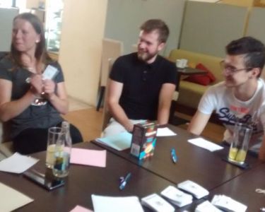 Group of people sitting at a table with drinks, pens, and The Well-being Game cards spread around.