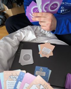 Person holding several The Well-being Game cards while sitting on a bed with more cards scattered on a table.